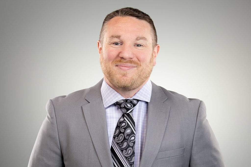 A man with short hair and a trimmed beard, wearing a light gray suit, patterned tie, and checked shirt, smiles at the camera against a plain gray background.