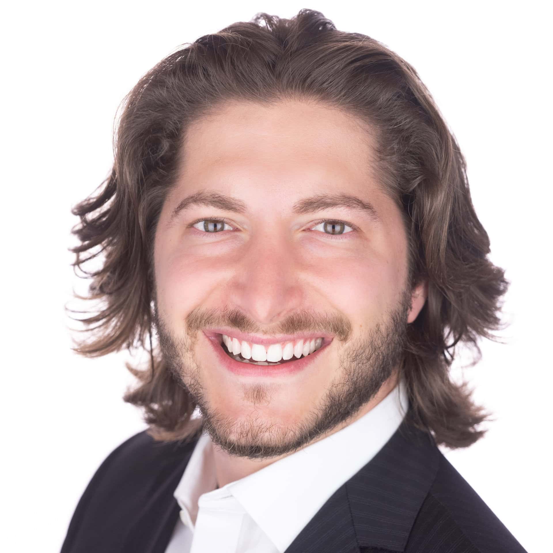 Smiling man with long brown hair and a beard, wearing a dark blazer and white shirt, against a plain white background.
