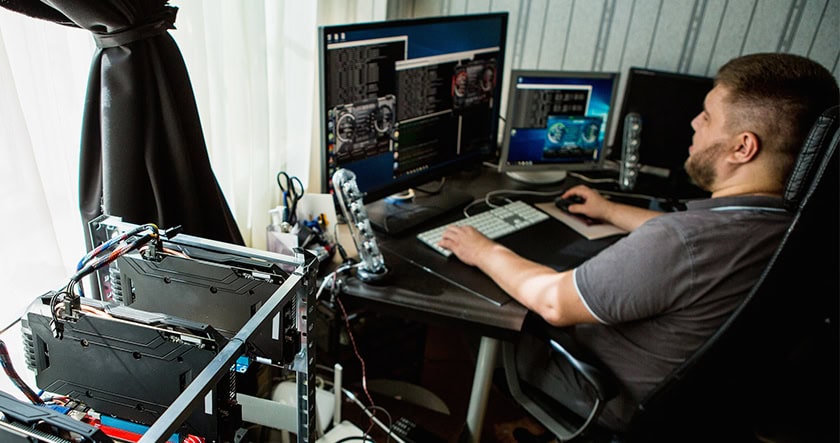 A man sits at a desk with multiple monitors, working on a computer. Nearby, there is cryptocurrency mining equipment beside the desk.