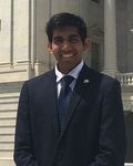 A person wearing a dark suit and tie stands outside in front of a stone building with columns, smiling at the camera.