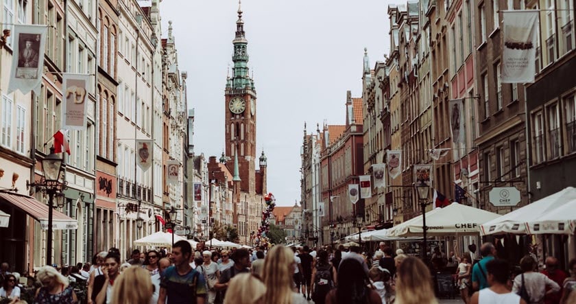 A crowded pedestrian street lined with historic buildings, outdoor cafes, and shops, leading to a tall clock tower in the distance under a cloudy sky.