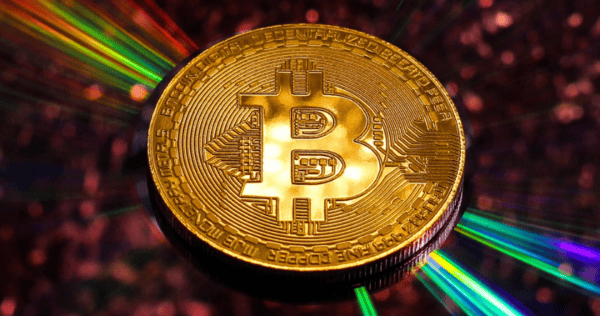 A gold Bitcoin coin with the Bitcoin symbol is shown in sharp focus, with colorful light streaks radiating from behind it against a blurred background.