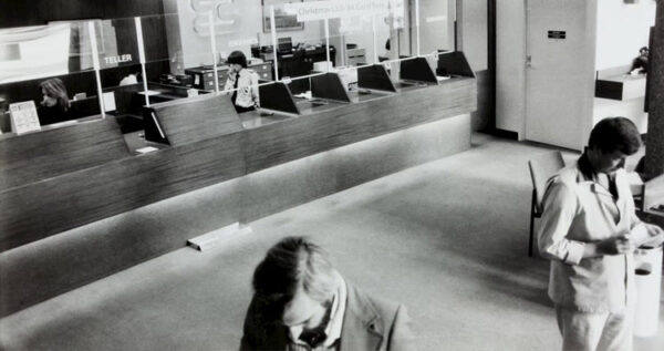 Black and white photo of a bank interior with tellers behind glass partitions and two male customers in the foreground.