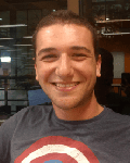 A young man with short dark hair smiles at the camera, wearing a Captain America T-shirt. The background shows an indoor office or workspace with glass walls.