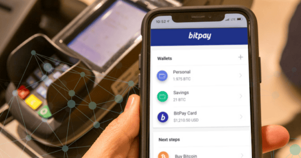 A person holds a smartphone displaying the BitPay app with wallet balances in front of a card payment terminal.