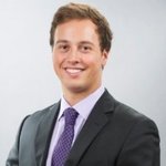 A young man in a business suit and tie smiles at the camera against a plain, light-colored background.