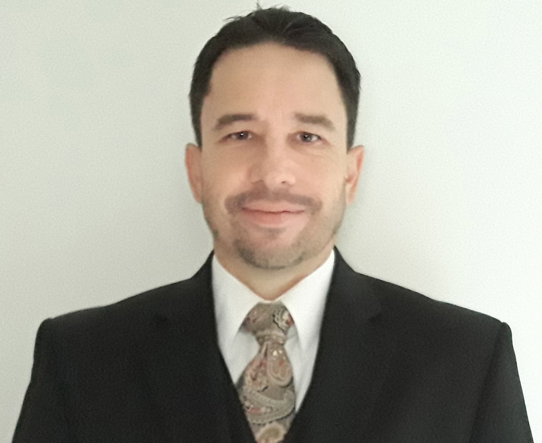 A man with short dark hair and a trimmed beard, wearing a black suit, white dress shirt, and a paisley-patterned tie, stands against a plain light-colored background and smiles slightly at the camera.