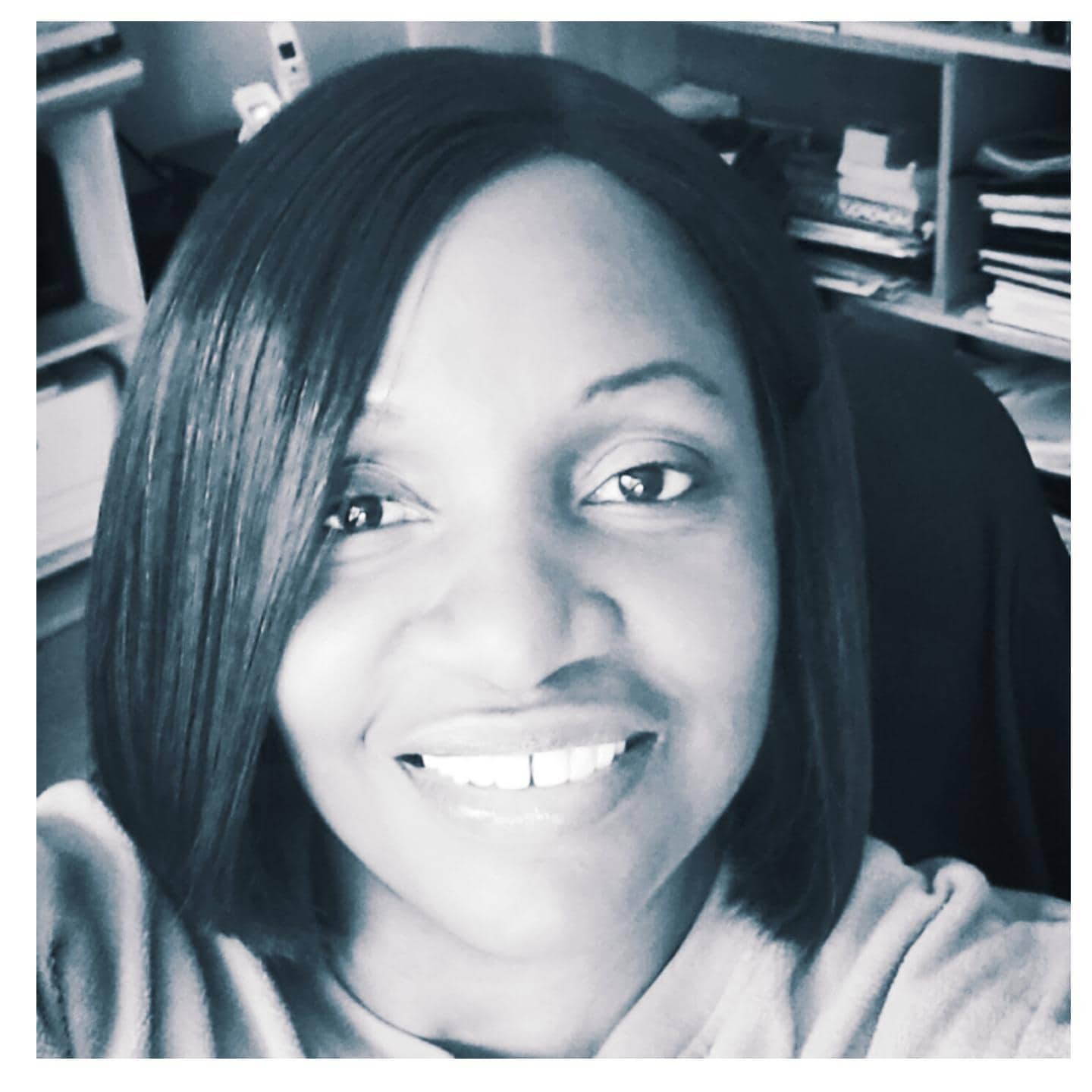 A woman with straight, shoulder-length hair smiles at the camera in a black and white close-up selfie, with shelves and stacked papers visible in the background.