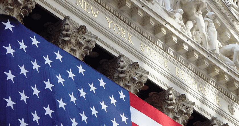 Large American flag hanging in front of the New York Stock Exchange building, with the building's name engraved above columns and decorative sculptures.
