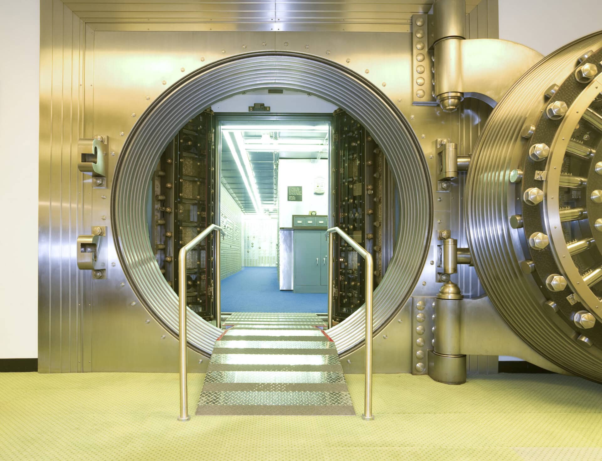 A large, circular metal bank vault door is open, revealing a brightly lit interior with blue flooring and metal walls. A metal ramp with railings leads into the secure vault room.