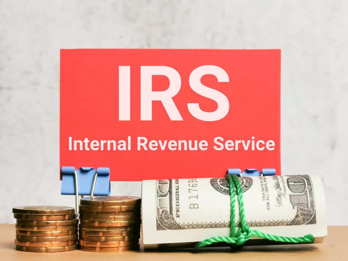 A red card reading IRS Internal Revenue Service stands behind stacked coins and a rolled-up bundle of U.S. hundred-dollar bills secured with a green string.