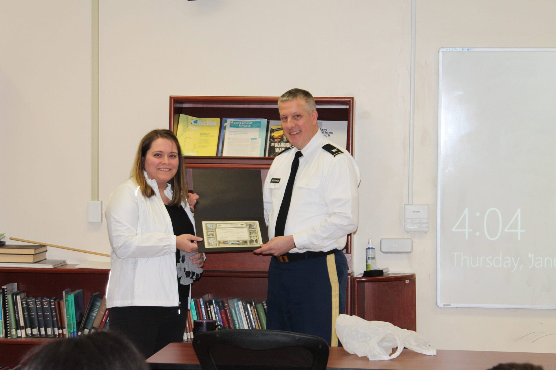 A woman and a man in a military uniform stand in an office, smiling, while the woman hands the man a certificate. Shelves with books and a digital clock are in the background.