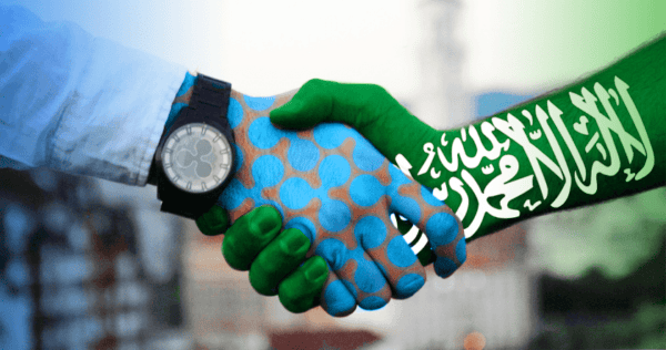 Two people shaking hands, one hand painted with the Saudi Arabian flag and the other painted with blue and green polka dots, symbolizing partnership or agreement.