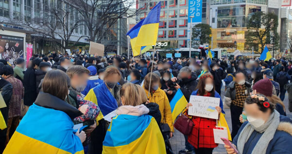 A large group of people gather outdoors, many holding or wearing Ukrainian flags and protest signs, in what appears to be a public demonstration.