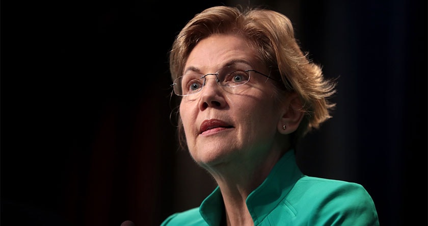 A woman with short blonde hair and glasses, wearing a green jacket, looks slightly upward while speaking in front of a dark background.