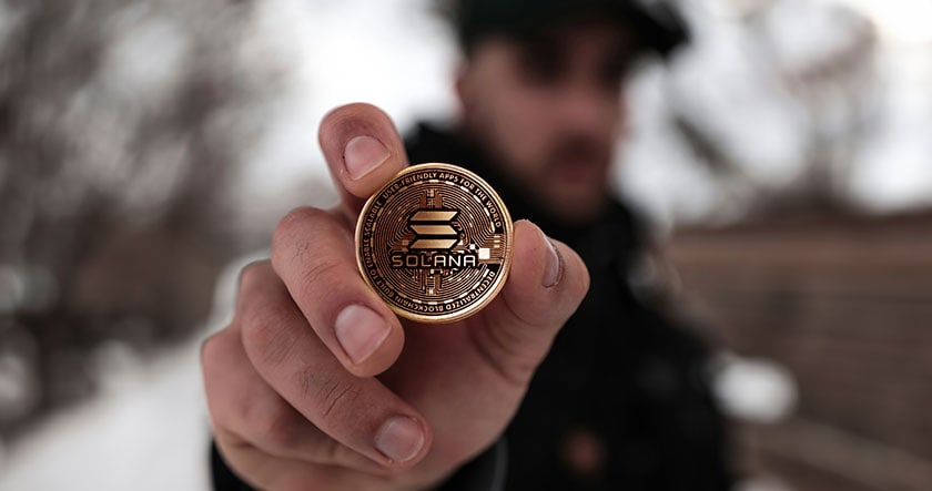A person holds a Solana cryptocurrency coin close to the camera, with a blurred outdoor background.