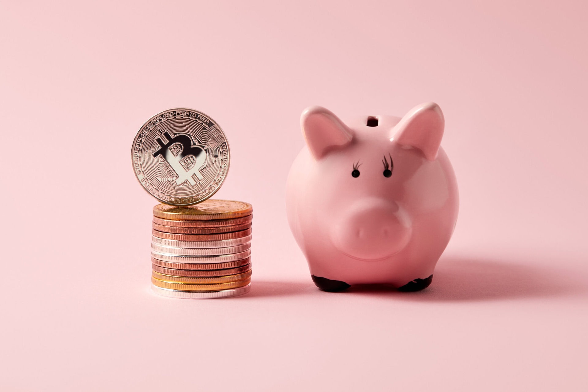 A pink piggy bank sits next to a stack of assorted coins topped with a silver Bitcoin, set against a soft pink background.