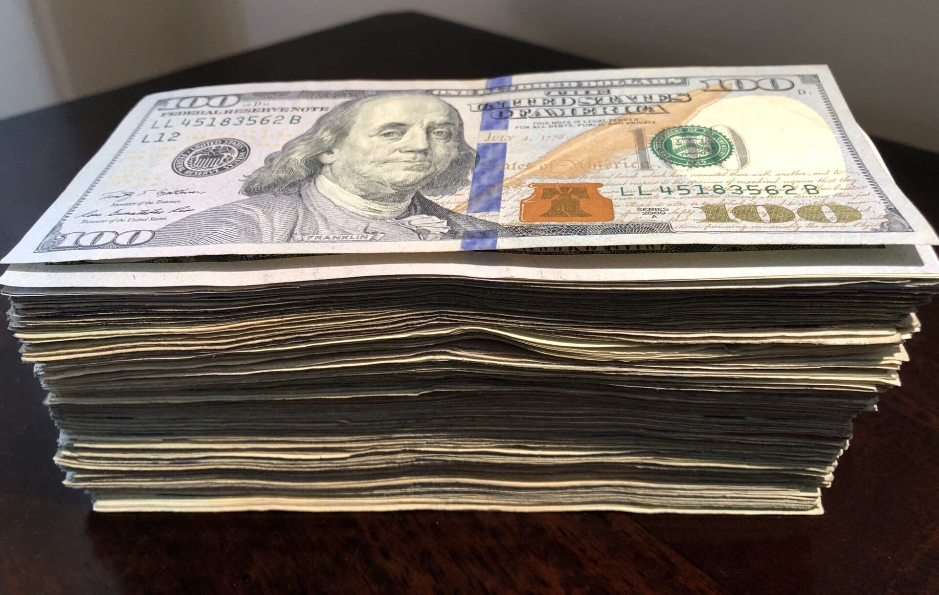 A large stack of U.S. $100 bills is placed on a dark wooden surface, with the top bill clearly showing Benjamin Franklins portrait.