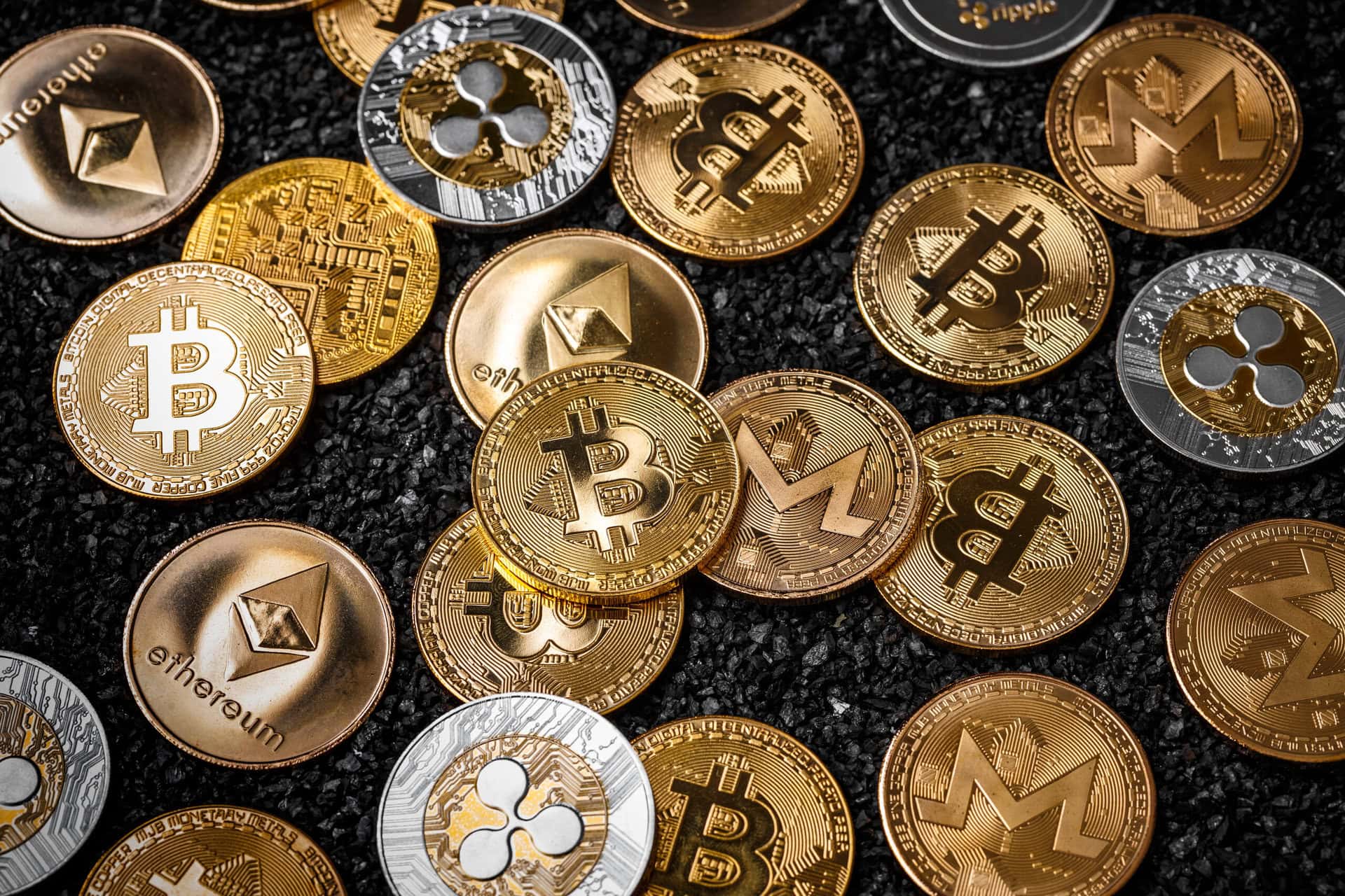 Assorted physical coins representing cryptocurrencies such as Bitcoin, Ethereum, Ripple, and Monero are scattered on a dark, textured surface.