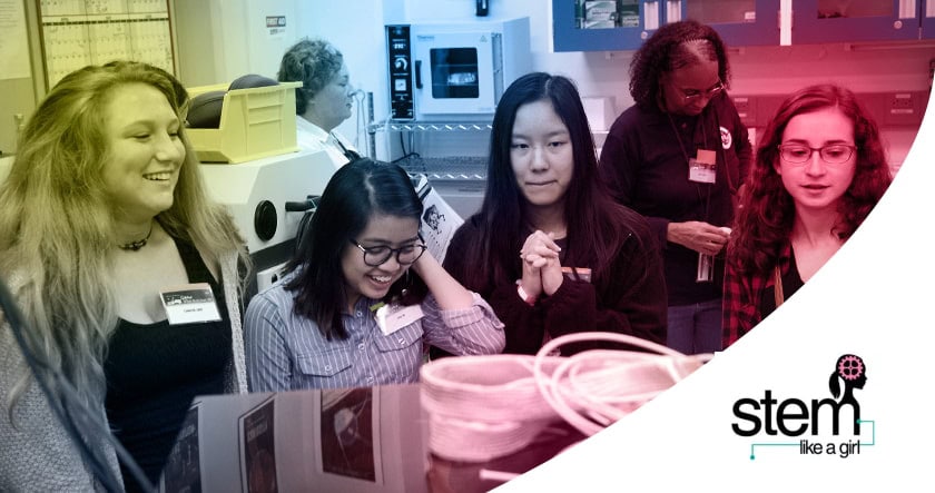Four young women observe a science demonstration in a lab setting, with the "STEM like a girl" logo in the bottom right corner.