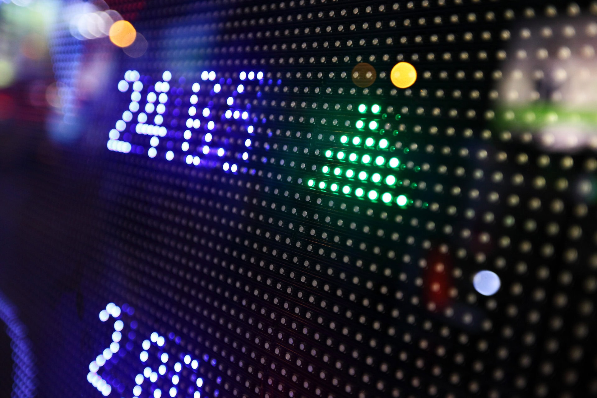 Close-up of an electronic stock market display showing numbers, with a green upward-pointing arrow indicating a rise in value. The background is blurred with colorful lights.