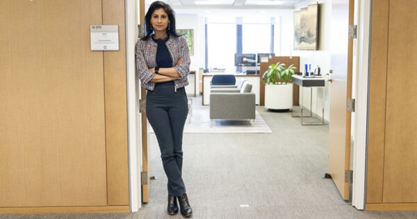 A woman stands with arms crossed in the doorway of a modern office, with a desk, chairs, and plants visible in the background.