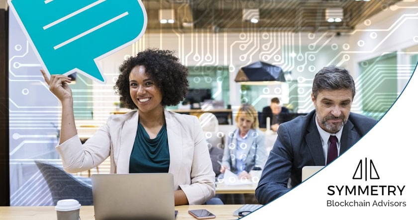 Two professionals sit at a desk with laptops; the woman is smiling and raising a chat symbol, while the man is focused. "Symmetry Blockchain Advisors" logo is visible.