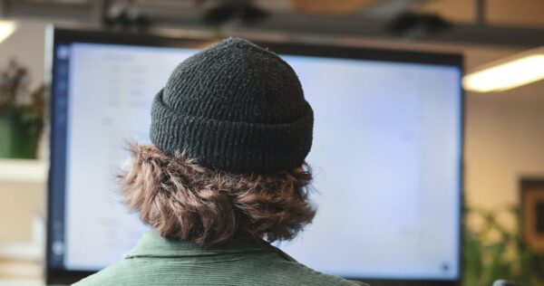 Person wearing a black knit beanie and green jacket is sitting and facing a large computer screen in an indoor setting.