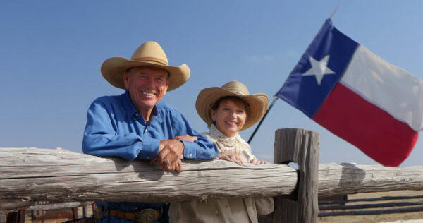 An older man and woman in cowboy hats lean on a wooden fence, smiling. A large Texas state flag waves in the background against a clear blue sky.