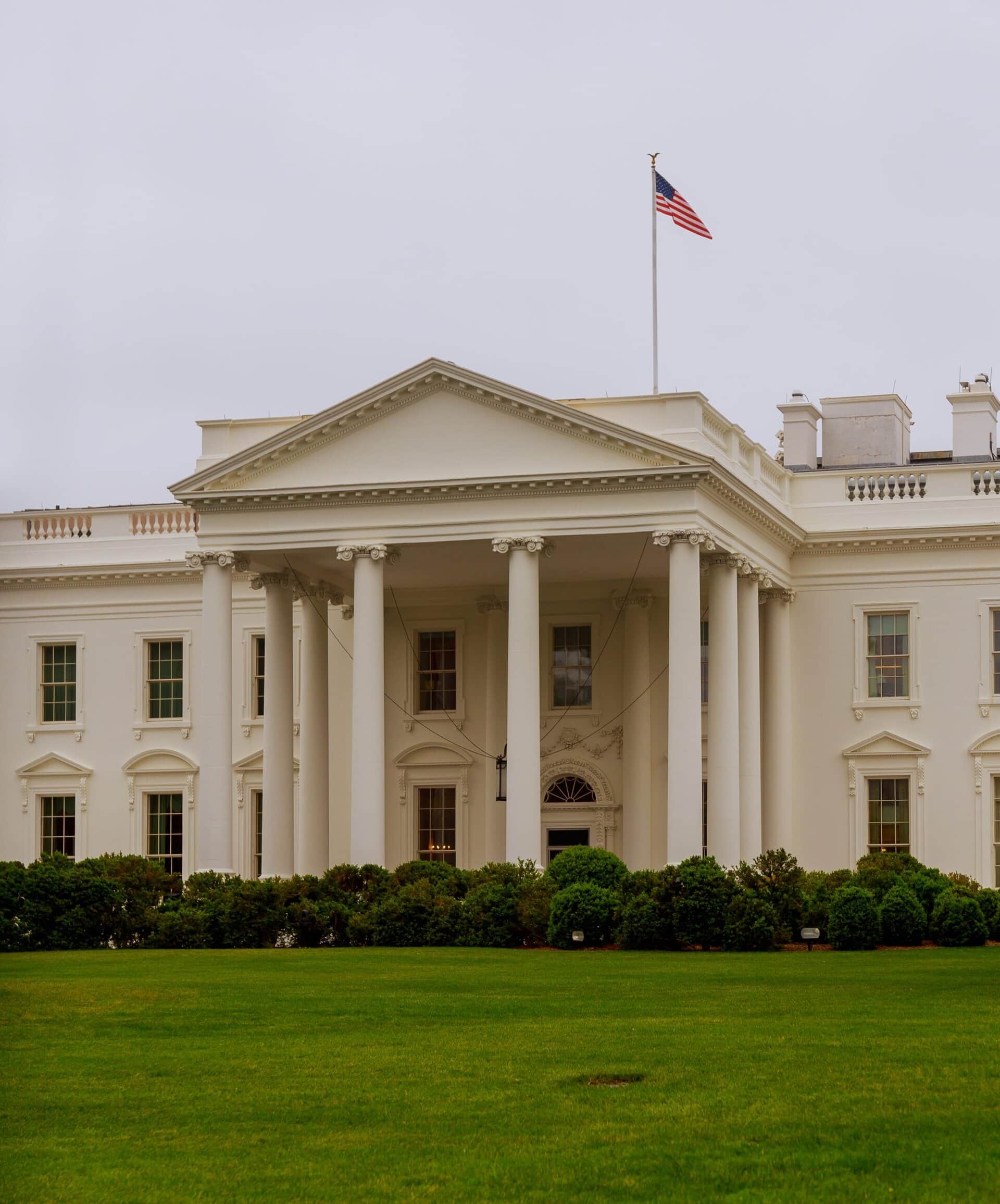 The White House with an American flag flying on the roof, large white columns at the entrance, and green lawn and bushes in the foreground under a cloudy sky.