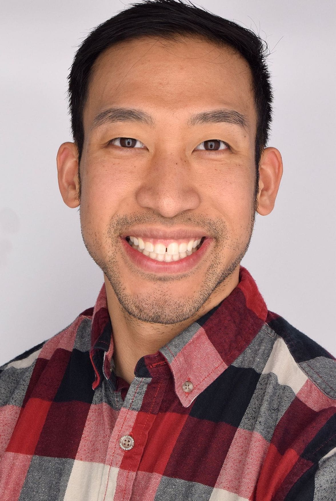 A man with short black hair and light facial hair smiles at the camera. He is wearing a red, black, and white plaid button-up shirt against a plain light background.