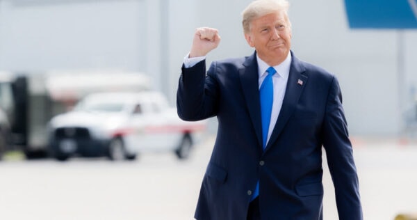 A man in a dark suit raises his fist while standing outdoors near a white vehicle.
