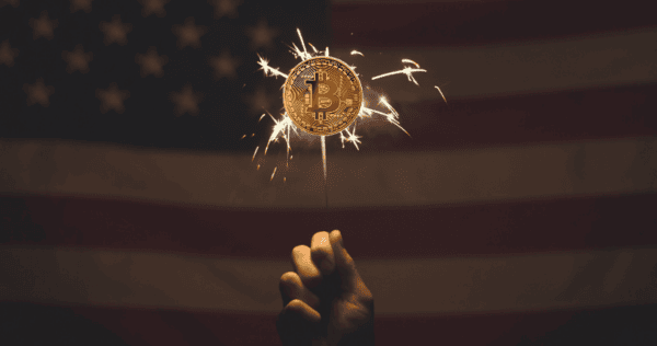A hand holds a sparkler with a Bitcoin symbol in front of a blurred American flag background.