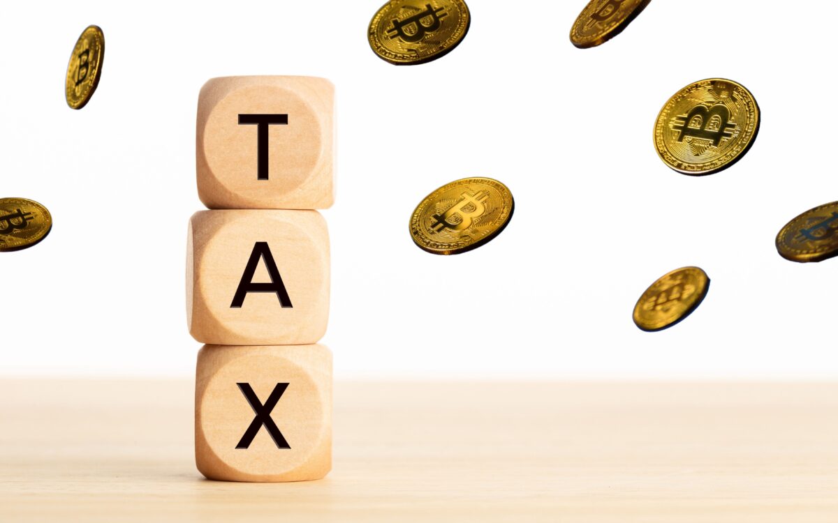 Three wooden blocks stacked to spell "TAX" with floating Bitcoin coins around them on a light wooden surface and white background.