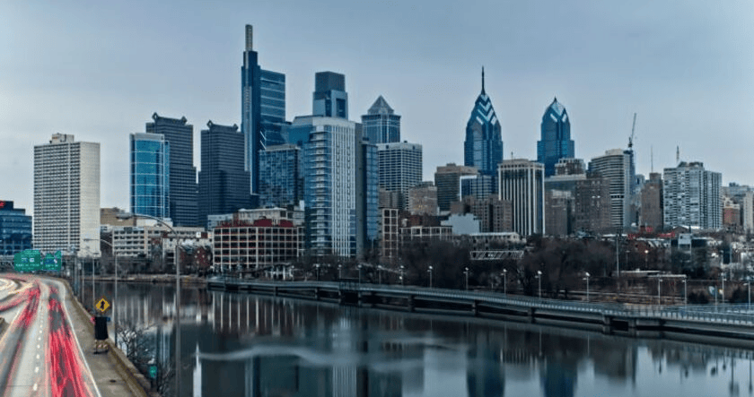 A city skyline with modern high-rise buildings and skyscrapers reflected in a river, with a highway on the left side of the image.