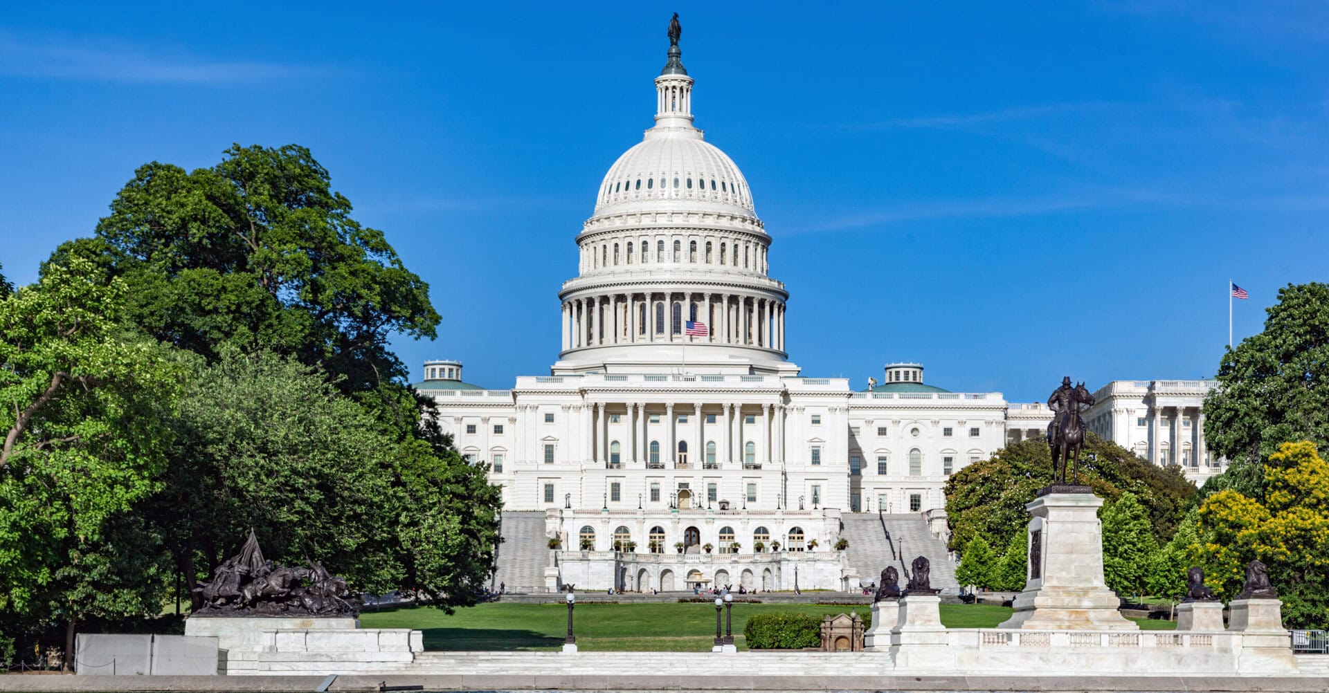 The United States Capitol building in Washington, D.C., features a large white dome with a statue on top, surrounded by trees and statues on a sunny day.