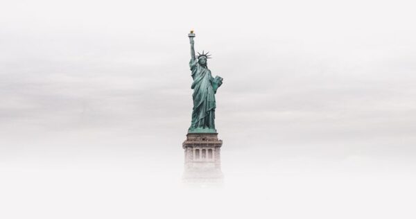 The Statue of Liberty stands tall against a cloudy sky, with its lower pedestal partially obscured by mist or fog.