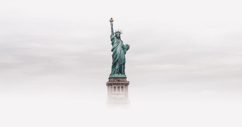The Statue of Liberty stands tall against a cloudy sky, with its lower pedestal partially obscured by mist or fog.