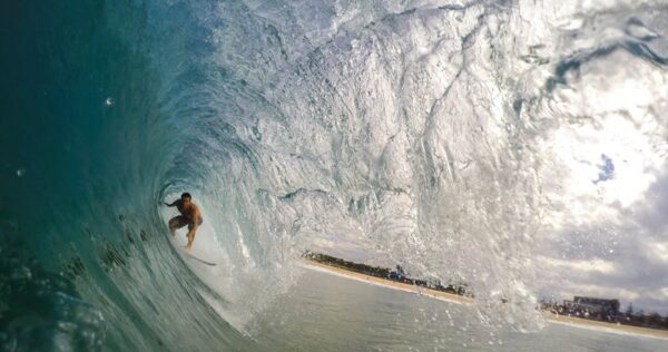 A surfer rides inside the barrel of a large, curling wave near the shoreline on a sunny day.