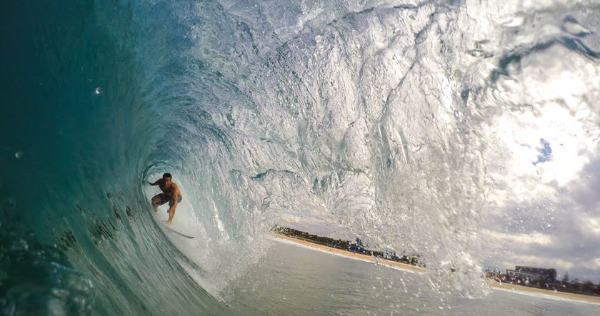 A surfer rides inside the barrel of a large, curling wave near the shoreline on a sunny day.