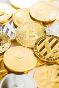 A close-up of various cryptocurrency coins, including Bitcoin, Ethereum, and Litecoin, stacked and scattered across a white surface. The coins are gold and silver, displaying prominent crypto logos and markings.