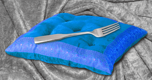 A metal fork resting on a blue velvet pillow with decorative trim, placed on a draped, gray velvet fabric background.