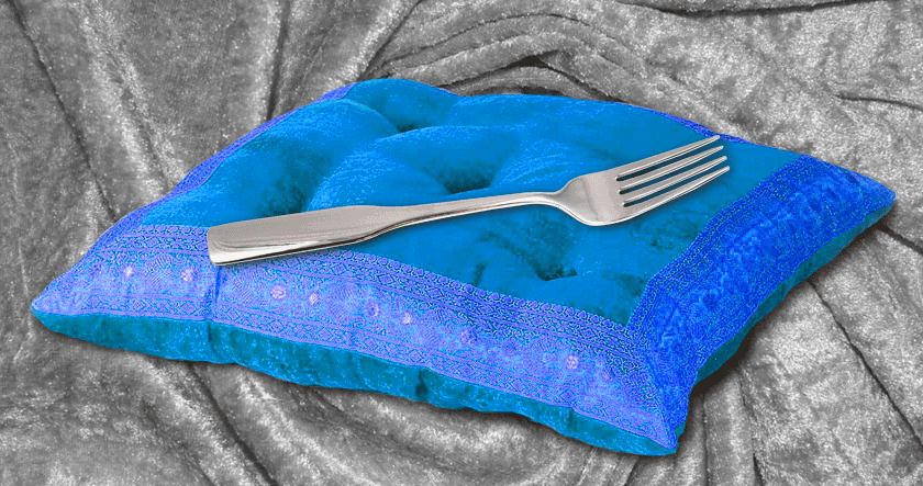 A metal fork resting on a blue velvet pillow with decorative trim, placed on a draped, gray velvet fabric background.