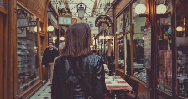 A person with shoulder-length hair and a black jacket walks through a narrow indoor shopping arcade lined with shops and warm lighting.