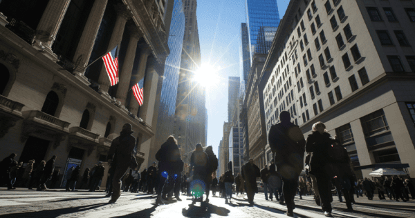 People walk along a busy city street in sunlight, with American flags on a building and tall skyscrapers in the background.