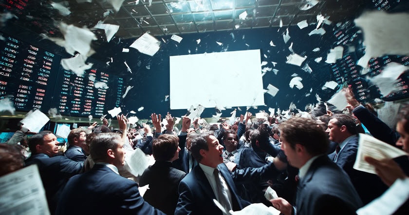 Crowded stock exchange trading floor with people in suits, tossing papers and raising hands, large digital boards and a blank central screen visible in the background.