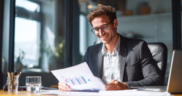 Man in a suit and glasses sits at a desk, smiling while reviewing printed charts and graphs, with a laptop, glass of water, and office supplies nearby.