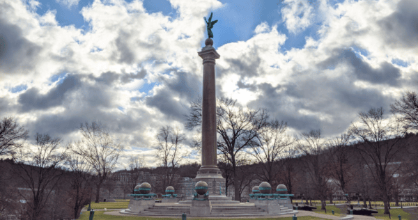 A tall column topped with a winged statue stands in a park, surrounded by smaller globe sculptures and leafless trees under a cloudy sky.