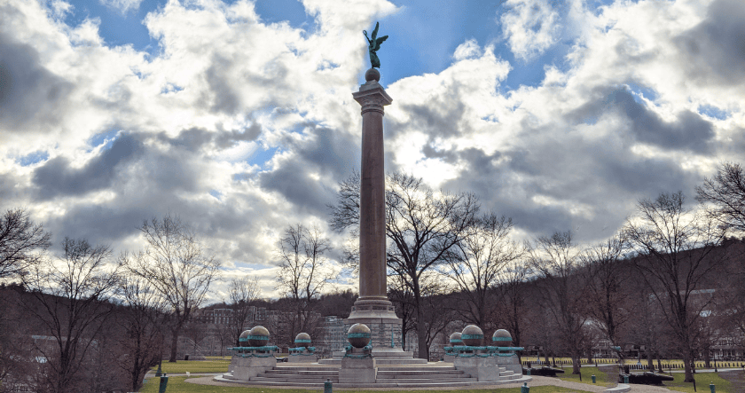 A tall column topped with a winged statue stands in a park, surrounded by smaller globe sculptures and leafless trees under a cloudy sky.