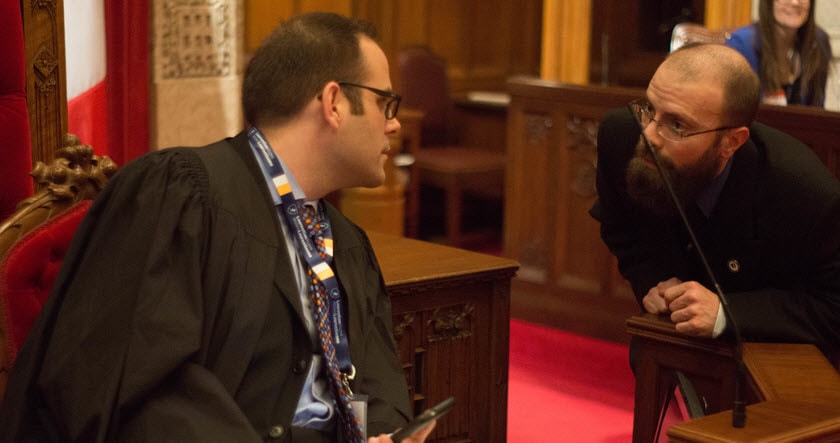Two men in formal attire have a discussion in a wood-paneled room with red carpet; one is seated while the other leans in to speak.